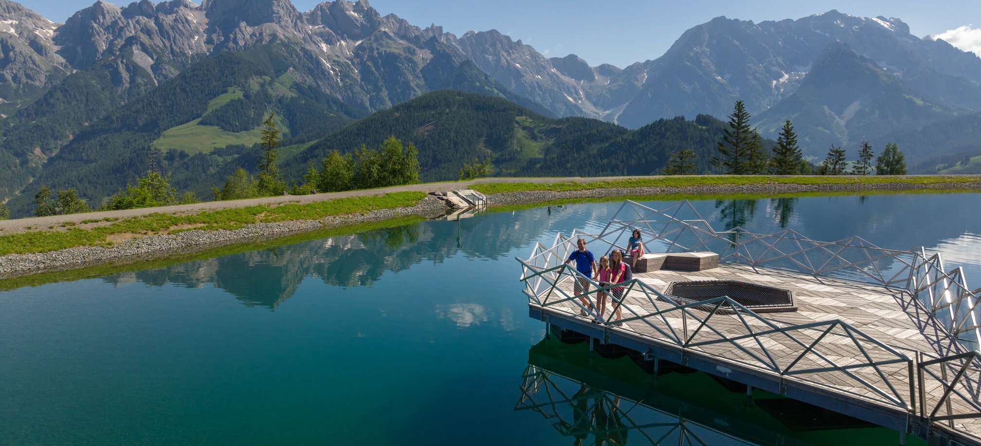 Family on wooden platform at Prinzensee in Maria Alm, clear lake water and Hochkönig mountains in background | © Roland Haschka