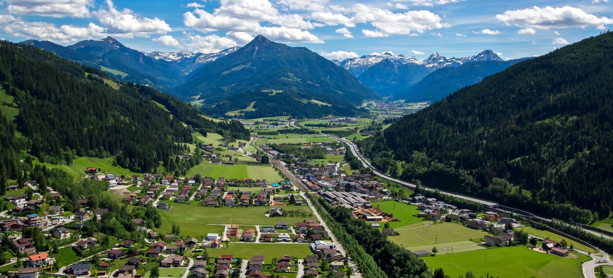 Panoramablick auf Eben im Pongau mit Wohnhäusern, Feldern, Autobahn und Bergen im Hintergrund an einem sonnigen Sommertag