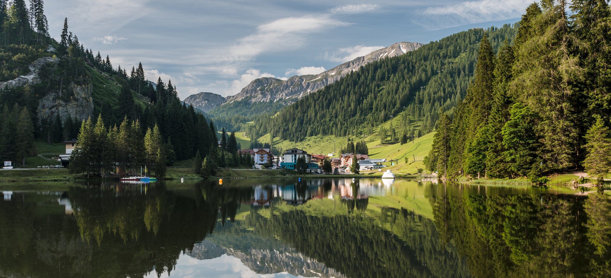 Mountain landscape with pine forest reflected in calm lake near Flachau, with boats, hotels and green alpine slopes. | © Matthias Fritzenwallner