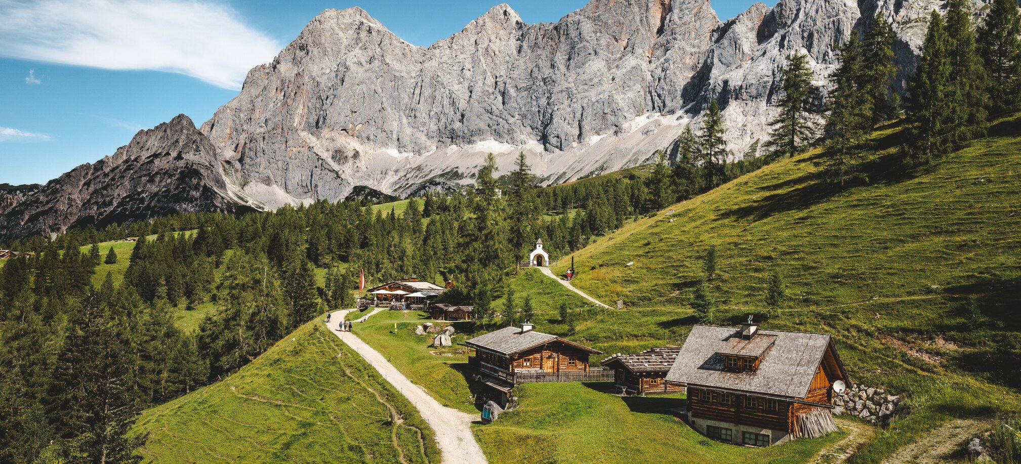 Wanderer auf einem Almweg bei urigen Hütten mit Kapelle, im Hintergrund das imposante Dachsteinmassiv bei blauem Himmel | © Gartner Mathäus
