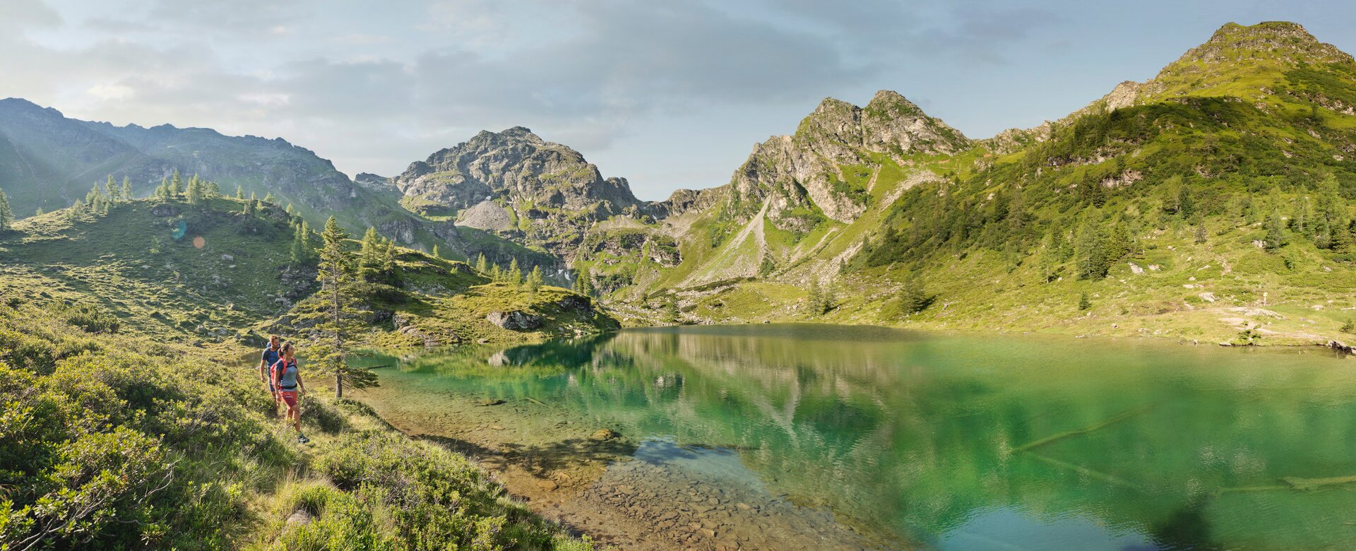 Two hikers walk along a turquoise mountain lake, surrounded by alpine meadows, trees and rugged mountain peaks | © Peter Burgstaller