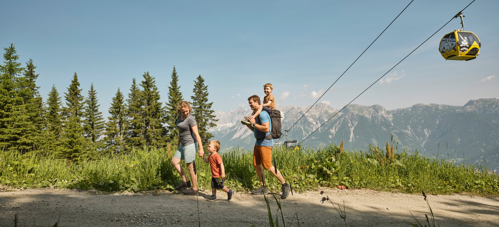 Family with two kids hiking on Planai with forest, mountains and a yellow cable car in the background | © Peter Burgstaller