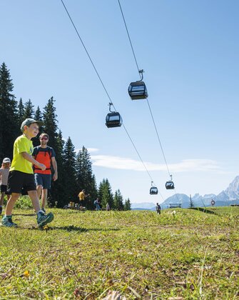 A family hikes across an alpine meadow beneath gondolas, enjoying the sunshine and mountain views in the Ski amadé region.