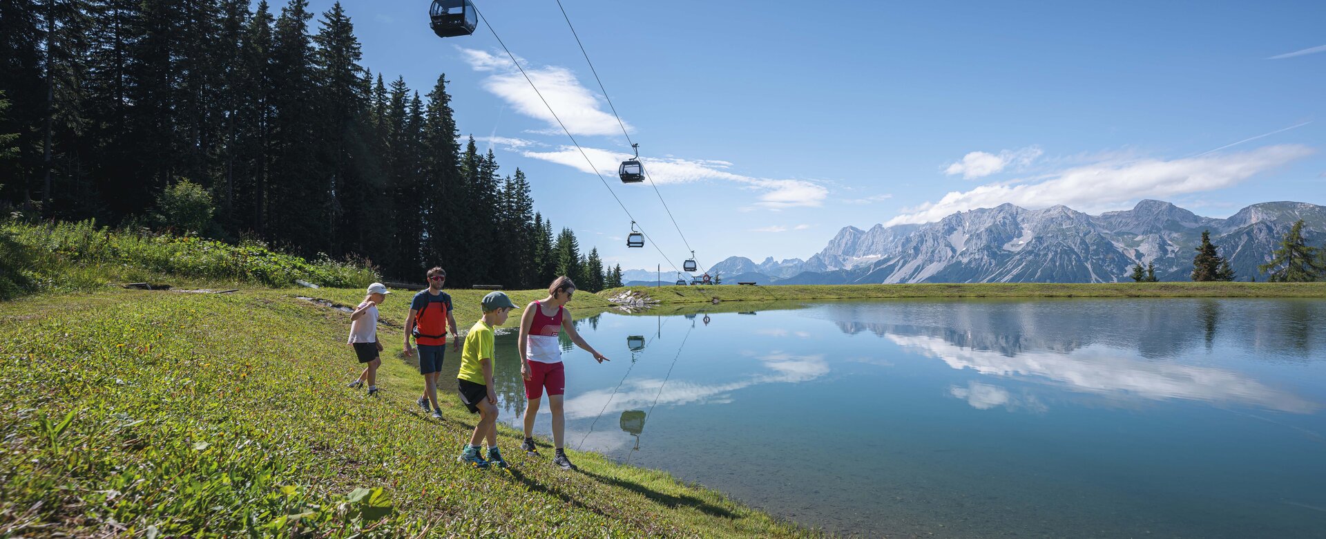 A family walks along a crystal-clear mountain lake under gondolas, enjoying the stunning view of the Dachstein mountains.