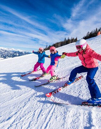 Three kids in colorful ski outfits ski side by side on the slope, holding hands and laughing in the snow.
