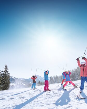 Kids in colorful ski outfits raise ski poles happily in the air, with snowy mountains and slopes in the background.