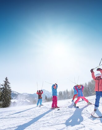 Kinder in bunter Skikleidung heben Skistöcke freudig in die Luft, vor verschneiten Bergen und Pisten.