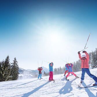 Kids in colorful ski outfits raise ski poles happily in the air, with snowy mountains and slopes in the background.