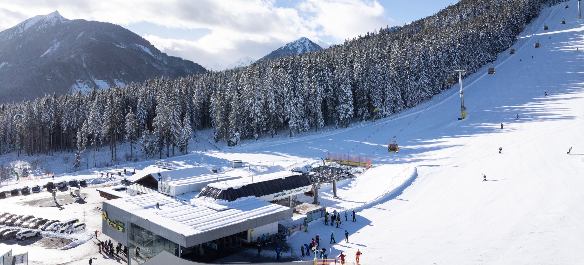 View of the valley station, skiers and gondolas, forest and mountains behind. | © Josh Absenger