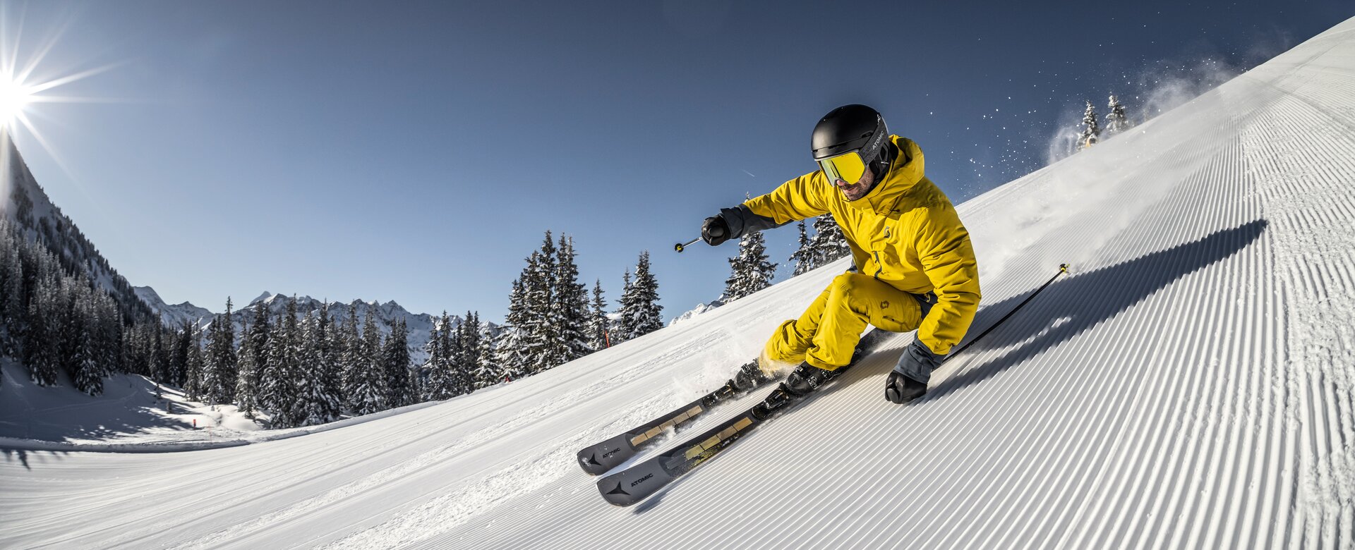 A skier in yellow outfit speeds down a freshly groomed slope, carving turns as snow sprays into the air. | © Mirja Geh