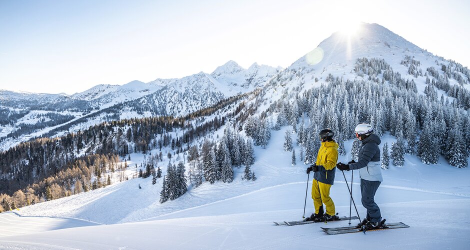 Two skiers in yellow and gray outfits stand on a freshly groomed slope, overlooking snow-covered mountains and forests. | © Mirja Geh
