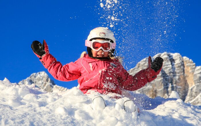 Child in pink ski gear playing happily in the snow with blue sky and mountains.