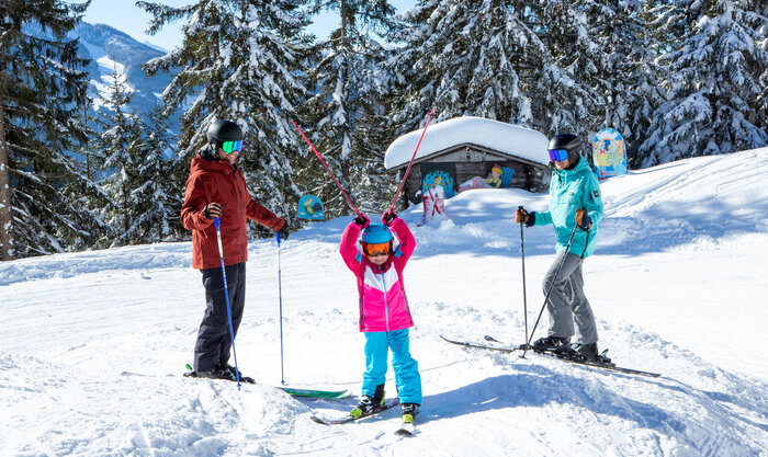 Family skiing on a snowy slope with winter figures and a hut in the background.