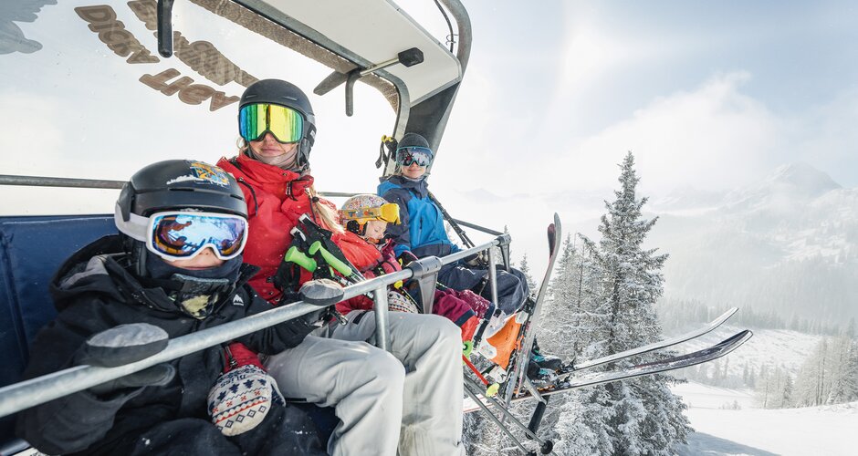 Family of four wearing ski gear on chairlift above snowy winter landscape with frosted trees | © Shuttleberg