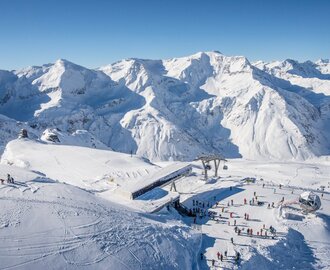  The top station of a cable car from above, with skiers next to it, surrounded by snow-covered mountains.