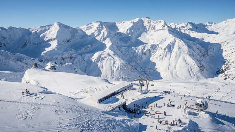  The top station of a cable car from above, with skiers next to it, surrounded by snow-covered mountains.