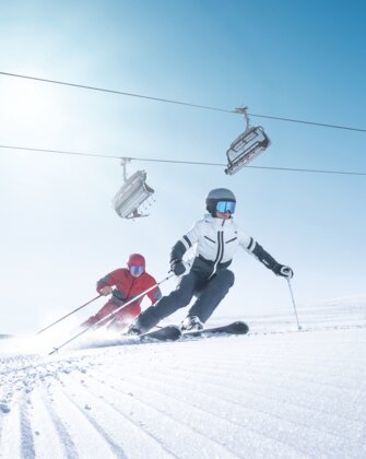 Two skiers carve in sync on freshly groomed slope in sunlight beneath operating chairlift