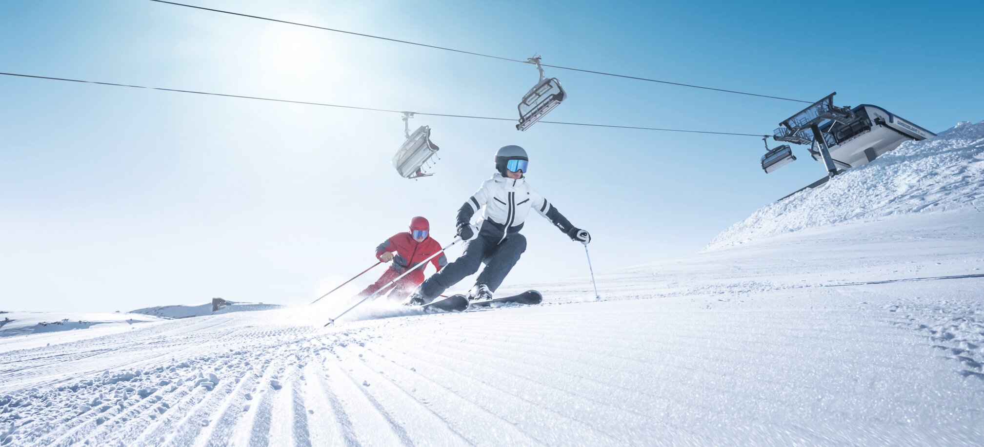 Zwei Skifahrer carven synchron auf frisch präparierter Piste bei Sonnenschein und Liftbetrieb