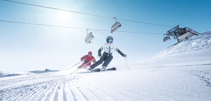 Two skiers carve in sync on freshly groomed slope in sunlight beneath operating chairlift