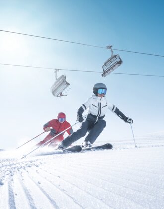 Zwei Skifahrer carven synchron auf frisch präparierter Piste bei Sonnenschein und Liftbetrieb