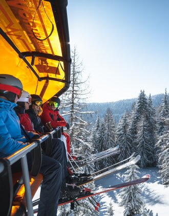 Four skiers wearing helmets and goggles sit on a chairlift with an orange cover above snow-covered trees. | © Ski amadé