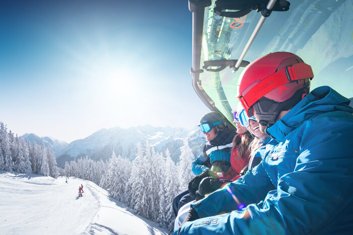 Group of skiers wearing helmets and goggles rides a chairlift with weather cover over a snow-covered mountain landscape. | © Ski amadé