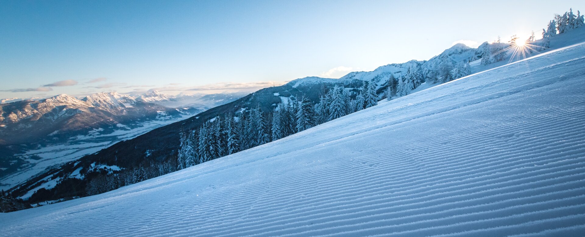 Groomed ski slope with sunrise over snowy mountains. | © Ski amadé