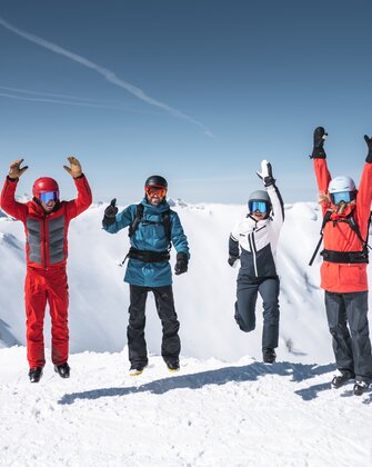 Four skiers jump in the air together with snowy mountain peaks and sunlight in the background