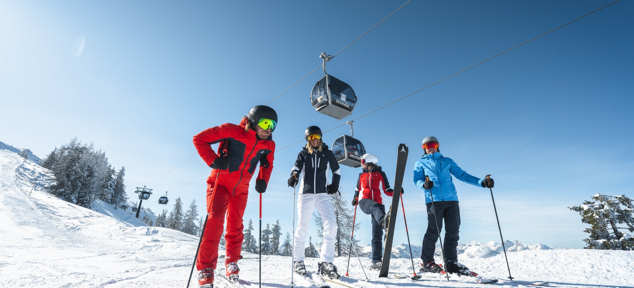 Skiers in colourful ski suits stand on the piste with the gondola hovering above them and one of them has lifted a ski. | © Ski amadé