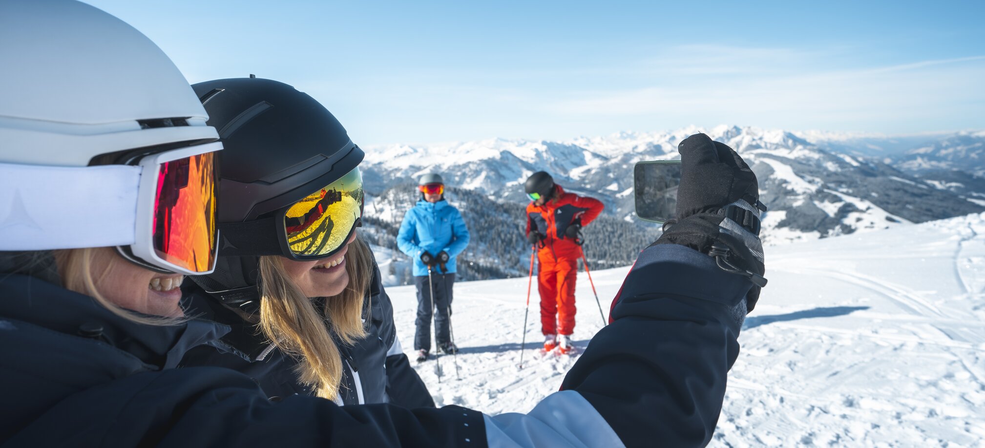 Two skiers take a selfie while two others pose in front of the snow-covered mountain landscape. | © Ski amadé