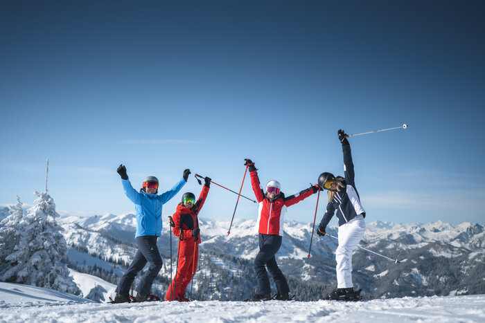 Four friends in ski equipment stand on the piste and stretch their arms joyfully in the air. | © Ski amadé