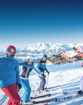 Four skiers laugh on the slope with a mountain hut in the background. | © Ski amadé