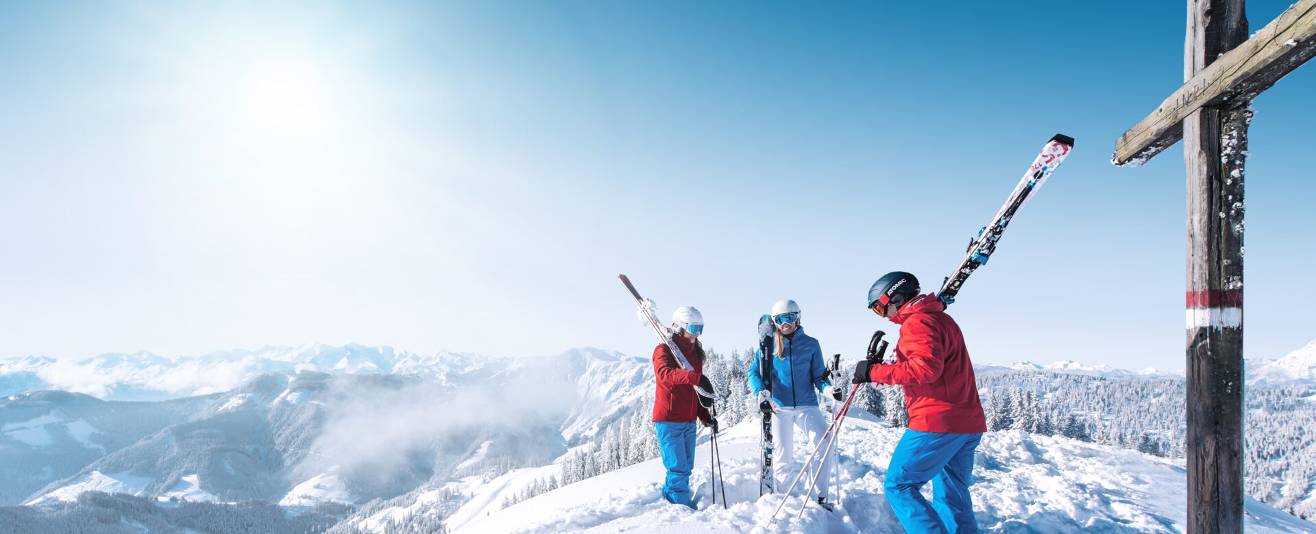 Three skiers stand by a summit cross, enjoying the mountain scenery. | © Ski amadé