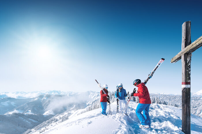 Three skiers stand by a summit cross, enjoying the mountain scenery. | © Ski amadé
