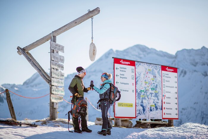 Two winter athletes stand by a weather station with a wooden sign in the mountains, examining a large ski trail map. | © Christoph Huber