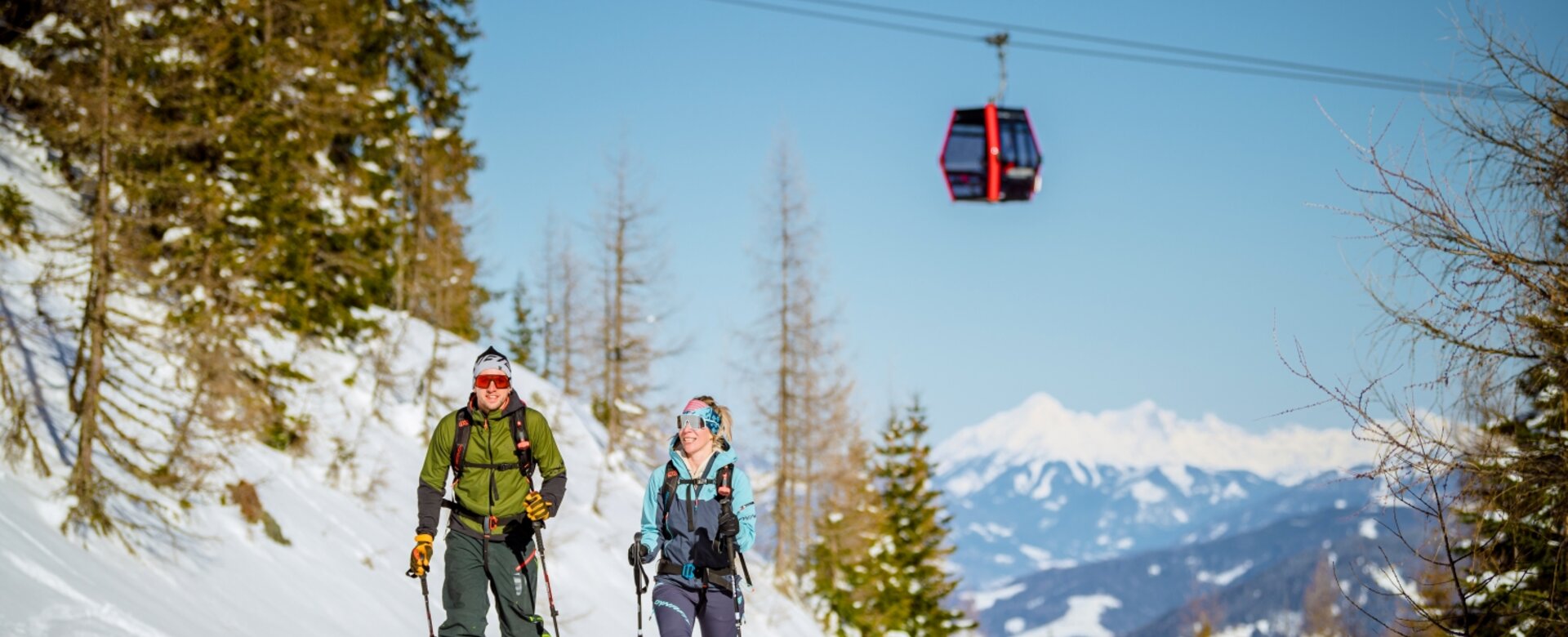 Two ski tourers ascend a snowy slope at Galsterberg, while a red cable car floats above them. | © Christoph Huber