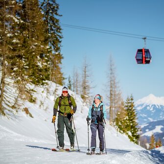 Two ski tourers ascend a snowy slope at Galsterberg, while a red cable car floats above them. | © Christoph Huber