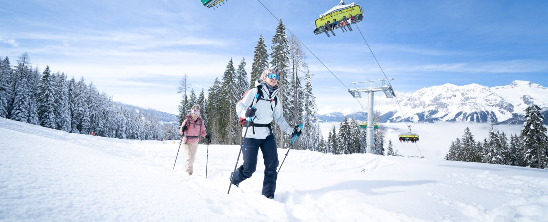 Two ski tourers ascend through the snow as a chairlift with skiers passes above them. | © Josh Absenger