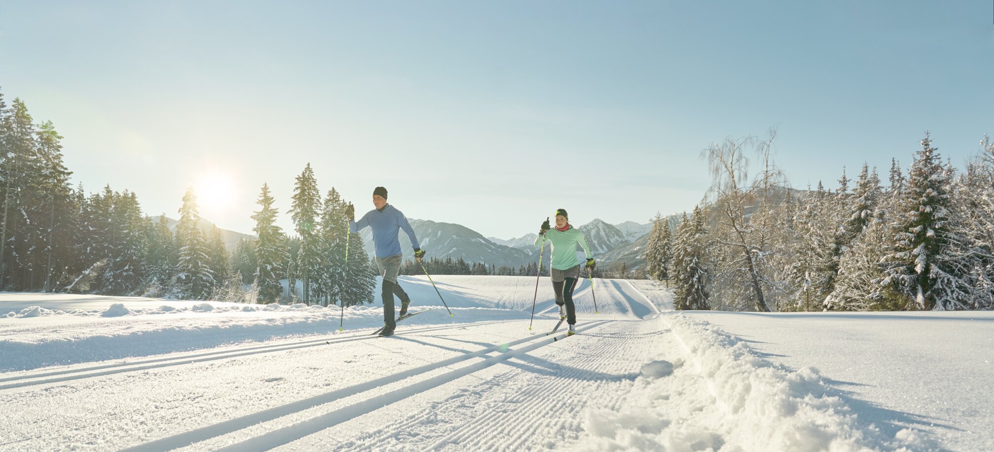 Two cross-country skiers in sportswear glide on sunny groomed trail through snowy winter landscape | © Peter Burgstaller