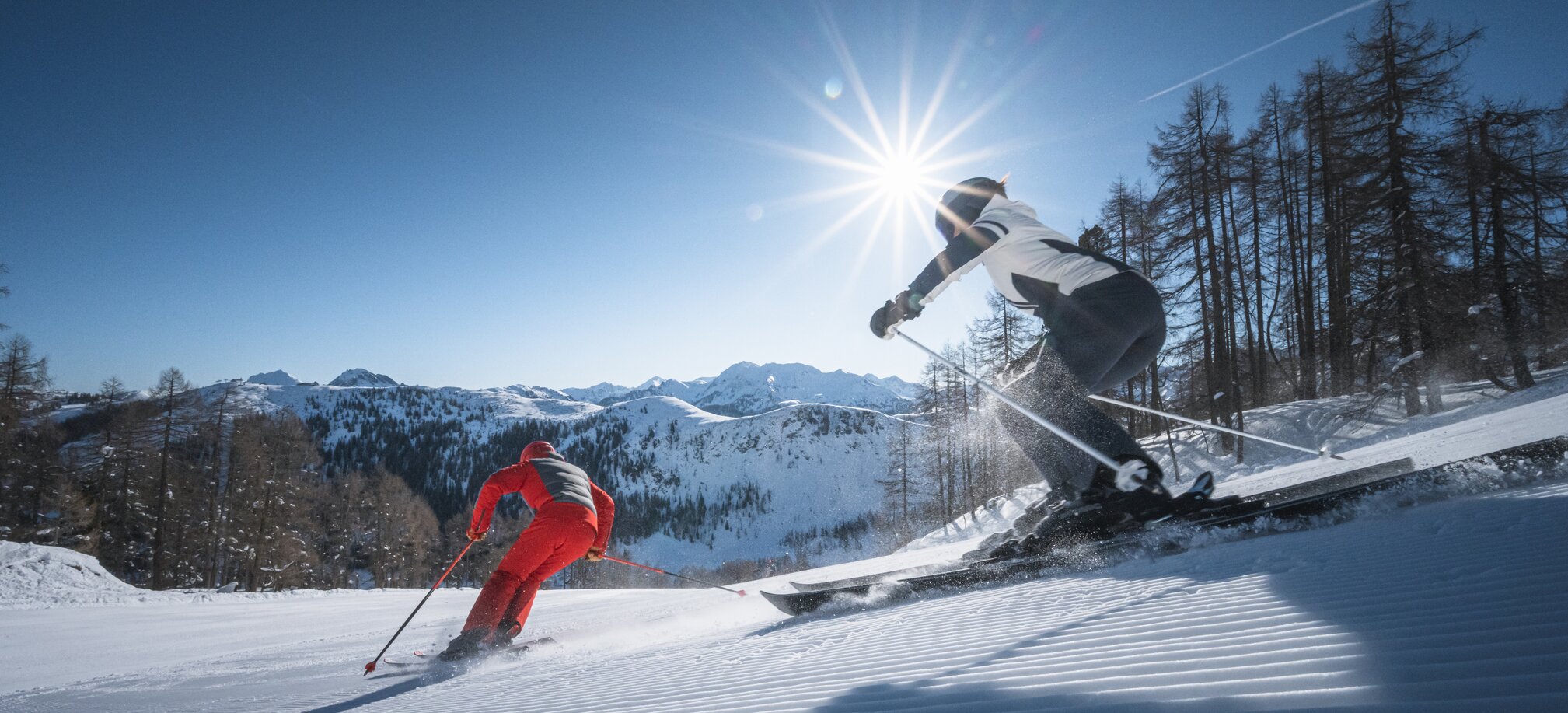 Two skiers carve down a freshly groomed slope, sunbeams shining through trees and above the mountain panorama.