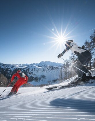 Zwei Skifahrer carven über frisch präparierte Piste, Sonnenstrahlen leuchten durch Bäume und über Berge.