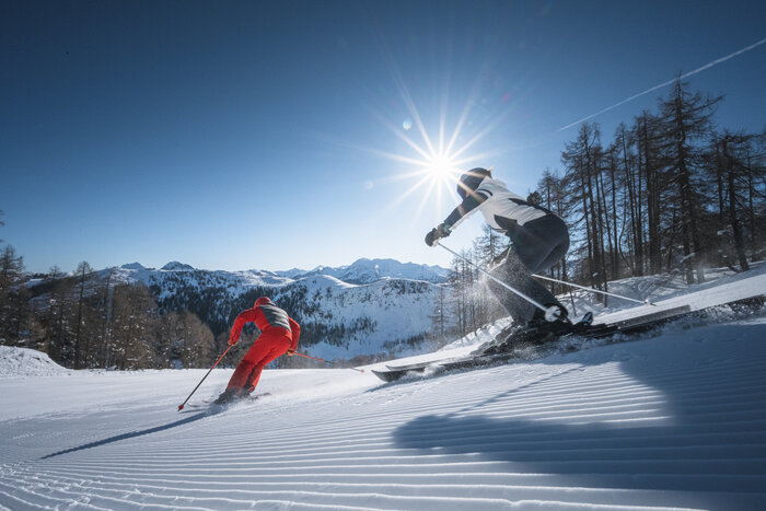 Two skiers carve down a freshly groomed slope, sunbeams shining through trees and above the mountain panorama.