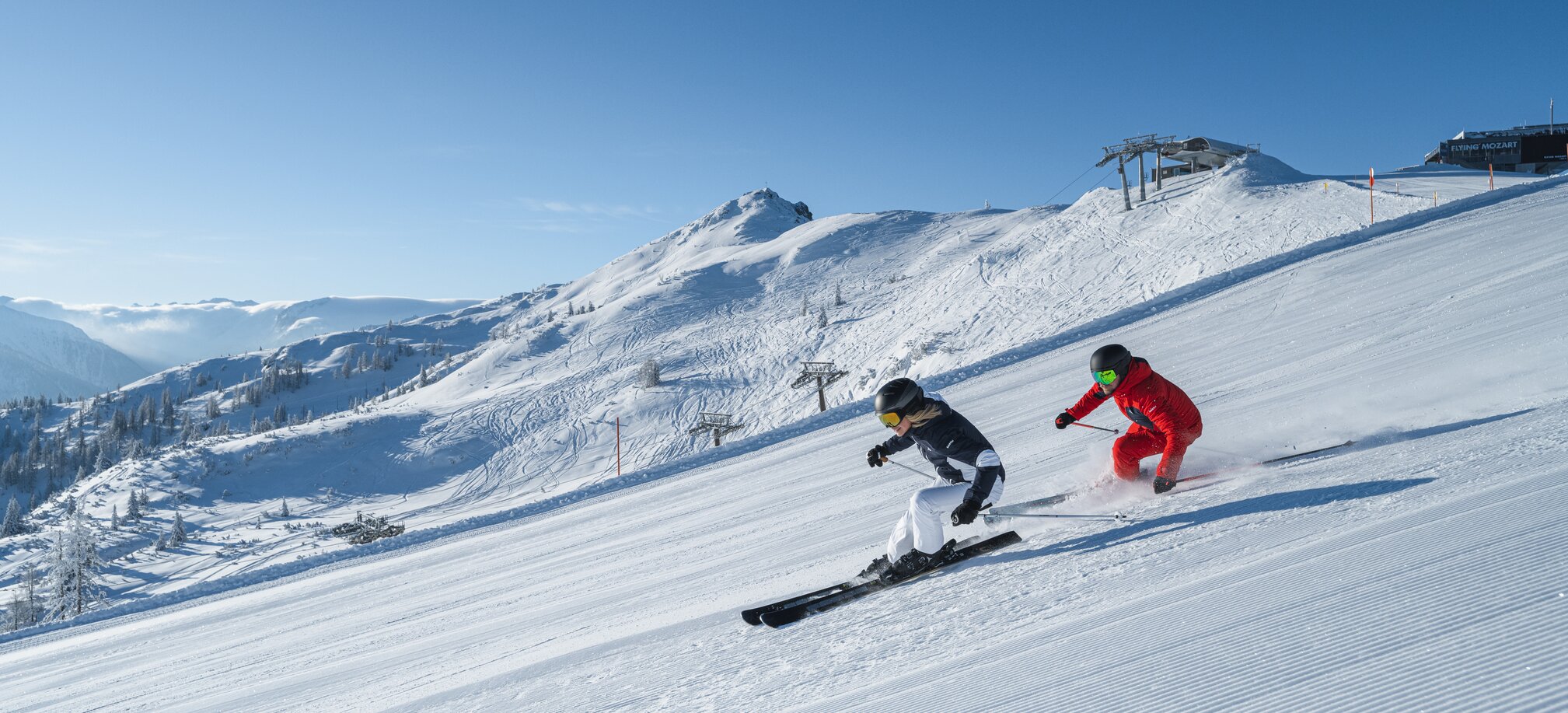 Zwei Skifahrer fahren parallel auf sonniger, gewalzter Piste mit Blick auf verschneite Berge und Sessellift