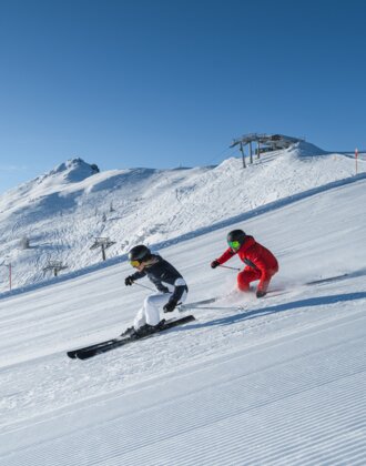 Zwei Skifahrer fahren parallel auf sonniger, gewalzter Piste mit Blick auf verschneite Berge und Sessellift
