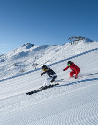 Zwei Skifahrer fahren parallel auf sonniger, gewalzter Piste mit Blick auf verschneite Berge und Sessellift