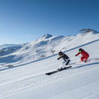 Zwei Skifahrer fahren parallel auf sonniger, gewalzter Piste mit Blick auf verschneite Berge und Sessellift