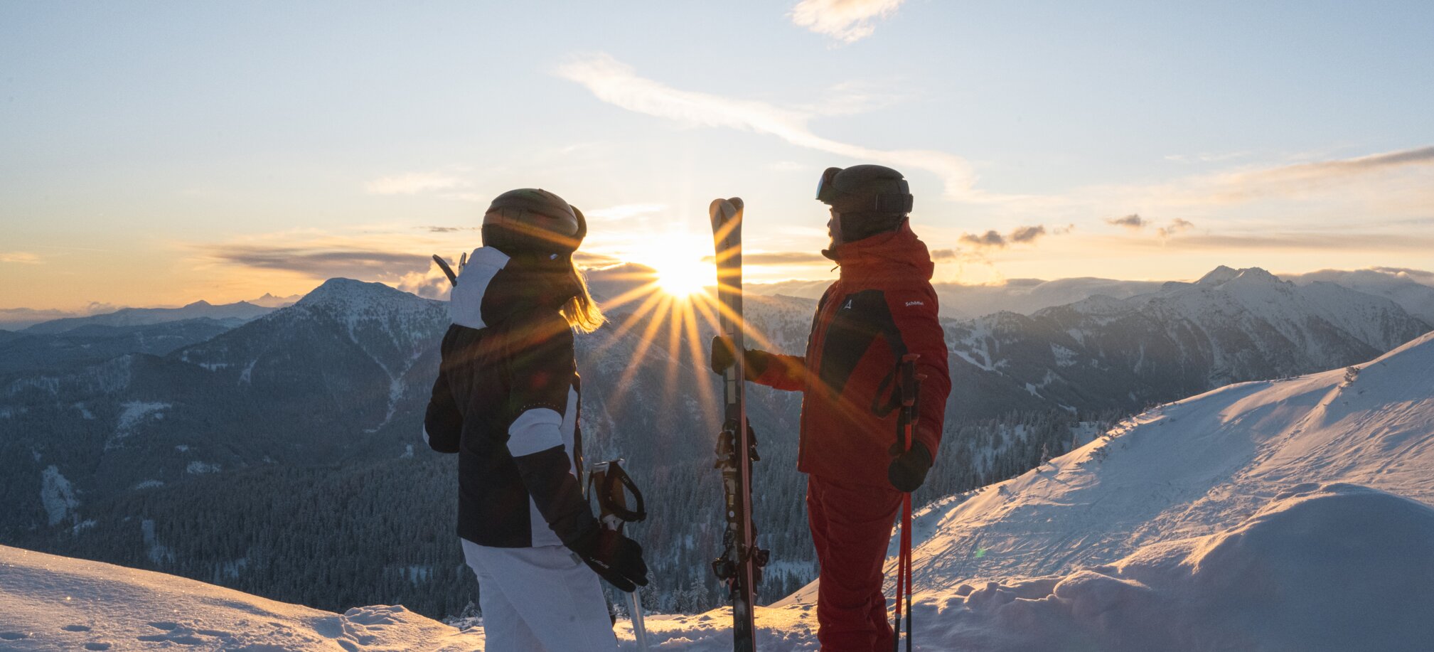 Two skiers in winter gear stand with skis on a snow-covered summit at sunrise in the mountains