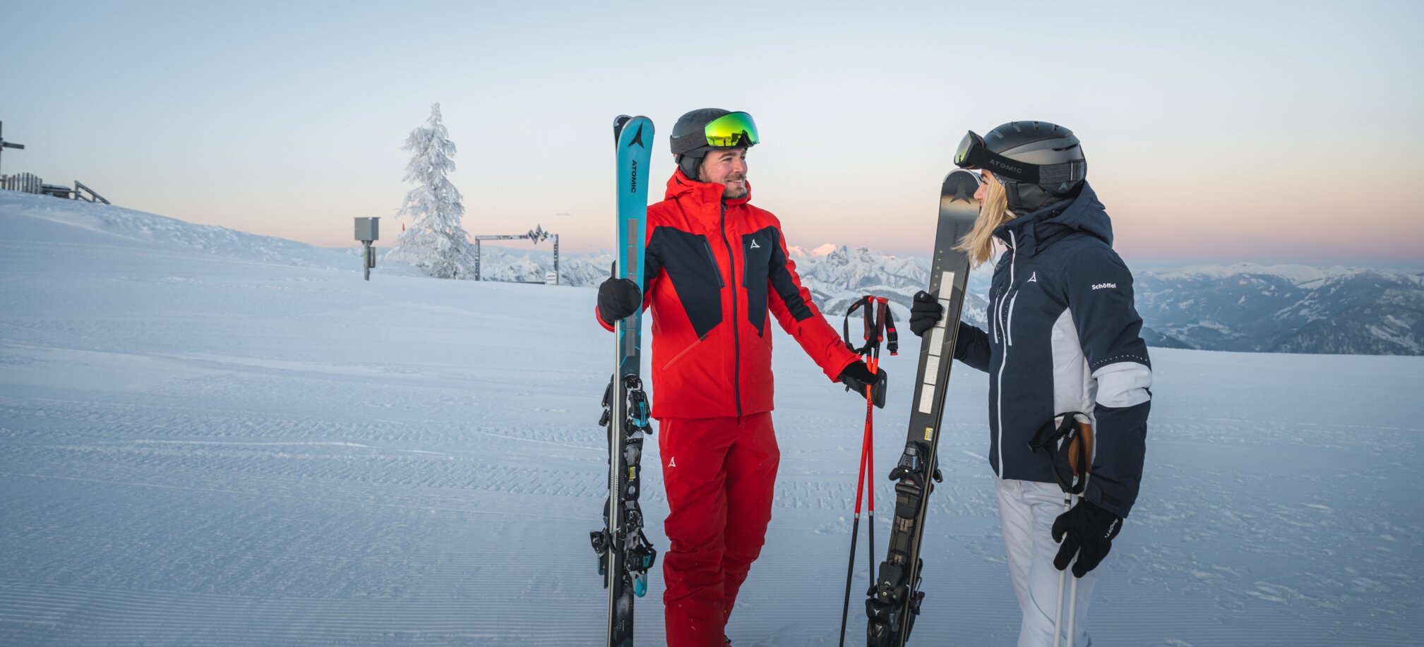 Ein Mann und eine Frau in Skikleidung stehen mit Skiern auf präparierter Piste vor Bergkulisse